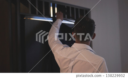 Rear view of a groom in a white dress shirt getting his suit jacket from a dark wardrobe. Rear view of a groom in a white dress shirt getting his suit jacket from a dark wardrobe. 138229932