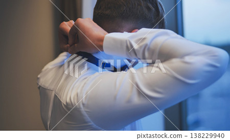 Groom getting ready for wedding, adjusting his blue bow tie in front of a window. Back view. 138229940