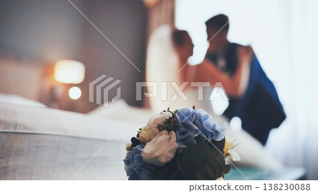 Blue and white wedding bouquet in the foreground with a blurred just married couple embracing in the background in their hotel room 138230088