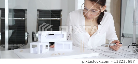 Female architect analyzing blueprints and a modern architectural model, working on new house design and construction planning in a professional office environment Female architect analyzing blueprints and a modern architectural model, working on new house design and construction planning in a professional office environment 138233697