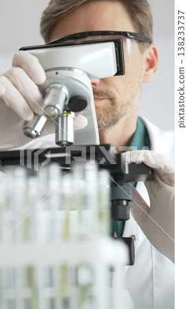 Man scientific researcher in protective gear analyzing specimens through a microscope in a sterile laboratory environment. Medicine, health care and science concept 138233737