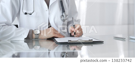 Female doctor in white coat with stethoscope writing patient notes on clipboard, completing medical records in clinic office. Medicine and health care 138233742