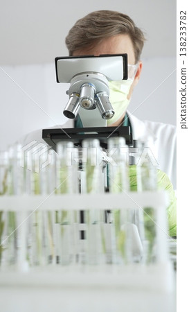Male scientific researcher in green protective gear analyzing specimens through a microscope in a sterile laboratory environment. Medicine and science concept 138233782