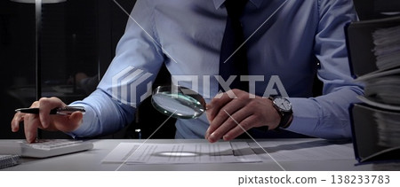 Financial auditor or accountant scrutinizing documents with a magnifying glass and calculator in a dark office. Audit and taxes in business Financial auditor or accountant scrutinizing documents with a magnifying glass and calculator in a dark office. Audit and taxes in business 138233783