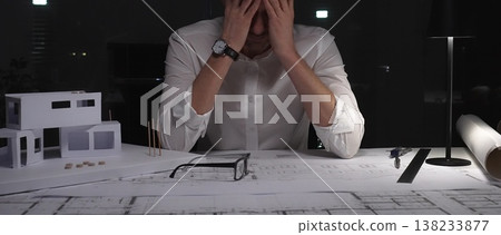 Architect experiencing burnout and stress, working late at night in a dark office with building models and blueprints. Architect experiencing burnout and stress, working late at night in a dark office with building models and blueprints. 138233877