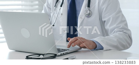 Medical doctor wearing lab coat and stethoscope is sitting at desk and typing on laptop, with eyeglasses and pen nearby, in bright medical office. Medicine and health care concept Medical doctor wearing lab coat and stethoscope is sitting at desk and typing on laptop, with eyeglasses and pen nearby, in bright medical office. Medicine and health care concept 138233883