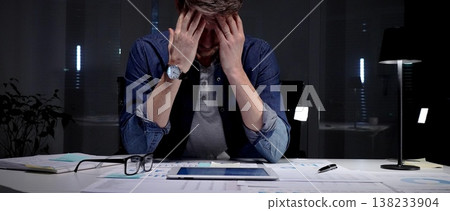 Young businessman with glasses sits at a dark office desk late at night, overwhelmed by documents and a tablet, clutching his head from stress, burnout and exhaustion 138233904