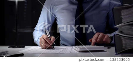 Businessman doing overtime work in office at night, using a digital tablet while surrounded by paper documents. Audit and taxes in business 138233905