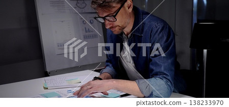 Professional man in glasses working late at night, analyzing financial data on a digital tablet, managing business reports, and extending productivity in a dark office environment Professional man in glasses working late at night, analyzing financial data on a digital tablet, managing business reports, and extending productivity in a dark office environment 138233970