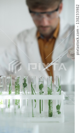 Man scientist wearing a lab coat, white gloves and protective glasses, is dripping liquid from a pipette into a test tube with a green plant inside, vertical portrait. Science and medicine concepts 138233982