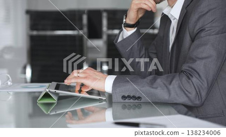 Business professional in a suit and wristwatch using a tablet on a glass desk, focused on data analysis and strategy planning in a modern corporate office environment Business professional in a suit and wristwatch using a tablet on a glass desk, focused on data analysis and strategy planning in a modern corporate office environment 138234006