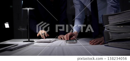 Business professionals collaborating over financial reports and a calculator beneath a desk lamp, reviewing documents and data late into the night to plan budget and strategy Business professionals collaborating over financial reports and a calculator beneath a desk lamp, reviewing documents and data late into the night to plan budget and strategy 138234056
