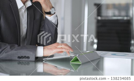 Business professional in a suit and wristwatch using a tablet on a glass desk, focused on data analysis and strategy planning in a modern corporate office environment Business professional in a suit and wristwatch using a tablet on a glass desk, focused on data analysis and strategy planning in a modern corporate office environment 138234085