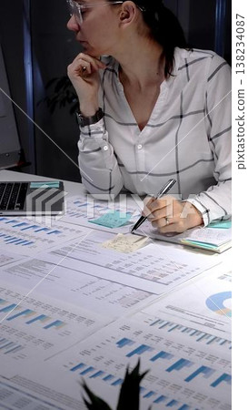 Businesswoman working late at a desk under a lamp, focused on documents and a tablet while finalizing reports and meeting a tight deadline in a corporate office setting Businesswoman working late at a desk under a lamp, focused on documents and a tablet while finalizing reports and meeting a tight deadline in a corporate office setting 138234087