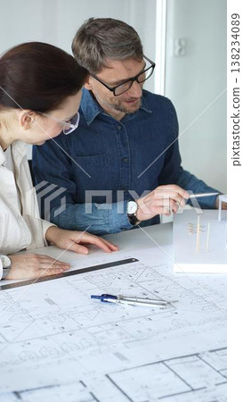 Architects and engineers collaborate over detailed blueprints and a house model, using compass and drafting tools to plan modern residential development with focused teamwork Architects and engineers collaborate over detailed blueprints and a house model, using compass and drafting tools to plan modern residential development with focused teamwork 138234089