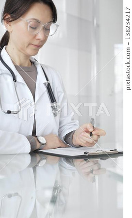 Female doctor in white coat and glasses writing medical notes on a clipboard at clinic desk, managing patient records. Medicine and health care Female doctor in white coat and glasses writing medical notes on a clipboard at clinic desk, managing patient records. Medicine and health care 138234217