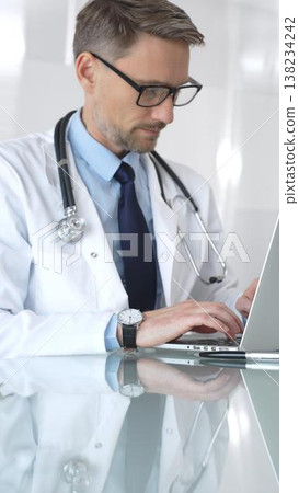 Male doctor in white coat with stethoscope, focused on laptop at reflective clinic desk, telemedicine consultation and research work. Medicine and health care Male doctor in white coat with stethoscope, focused on laptop at reflective clinic desk, telemedicine consultation and research work. Medicine and health care 138234242