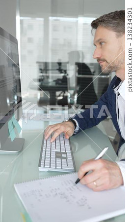 Professional businessman in a blue jacket working on desktop computer in modern office, focusing on his work and business tasks. Business people concept 138234301