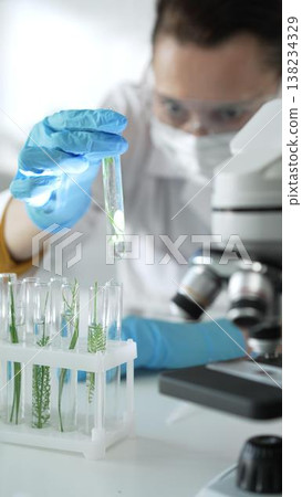 Woman scientist with blue gloves, face mask and protective glasses, is holding a test tube with plants inside near microscope in laboratory, vertical portrait view. Science and medicine concept Woman scientist with blue gloves, face mask and protective glasses, is holding a test tube with plants inside near microscope in laboratory, vertical portrait view. Science and medicine concept 138234329