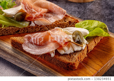 Rustic rye bread toasts with cured ham, cheese, and lettuce on a wooden cutting board. Rustic rye bread toasts with cured ham, cheese, and lettuce on a wooden cutting board. 138234931