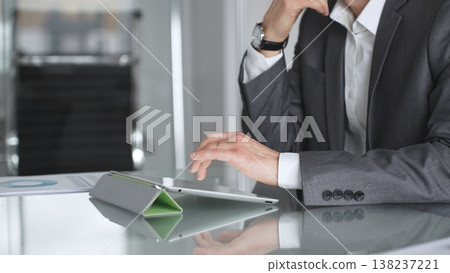Business professional in a suit and wristwatch using a tablet on a glass desk, focused on data analysis and strategy planning in a modern corporate office environment Business professional in a suit and wristwatch using a tablet on a glass desk, focused on data analysis and strategy planning in a modern corporate office environment 138237221