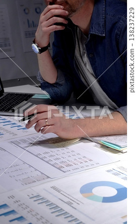 Caucasian man sitting at an office desk in the evening, concentrating and writing notes while surrounded by business documents, charts, graphs, and a laptop Caucasian man sitting at an office desk in the evening, concentrating and writing notes while surrounded by business documents, charts, graphs, and a laptop 138237229