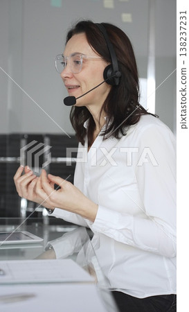 Businesswoman in a white shirt and headset leading a video call from a modern office, presenting with a tablet and gesturing as she discusses strategy and trains remote colleagues 138237231