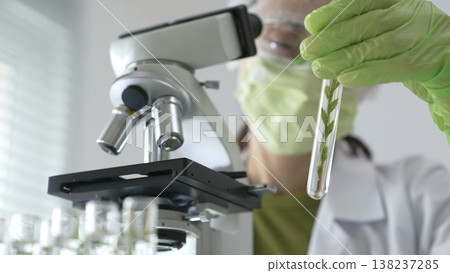 Female scientist wearing green protective mask and gloves is holding test tube with plant sample, researching botany and medical applications using a microscope. Medicine and science concept 138237285
