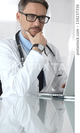 Male doctor wearing spectacles, thinking about healthcare diagnosis while using a laptop in a clinic office. Medicine and health care Male doctor wearing spectacles, thinking about healthcare diagnosis while using a laptop in a clinic office. Medicine and health care 138237356