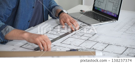 Architect sketching building plans on paper using a ruler and pen while working digitally on a laptop with a blueprint software displaying a floor plan Architect sketching building plans on paper using a ruler and pen while working digitally on a laptop with a blueprint software displaying a floor plan 138237374