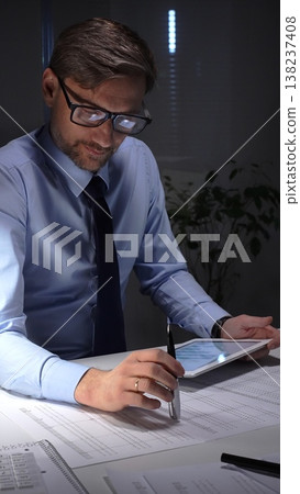 Businessman working late analyzing financial data at office, holding tablet with charts, writing notes on documents. Audit and taxes in business 138237408