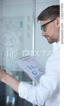 Young professional in glasses mapping business strategy and analyzing charts on a glass whiteboard, holding a clipboard while planning project goals, growth and execution 138237409