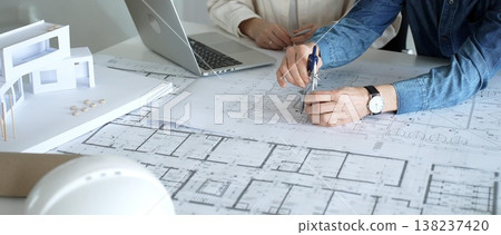 Engineering professionals collaborating on an architectural blueprint, using a compass to design a modern building, planning construction with a house model and laptop Engineering professionals collaborating on an architectural blueprint, using a compass to design a modern building, planning construction with a house model and laptop 138237420