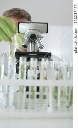 Man scientist wearing green protective gloves and mask is looking at green plant in test tube near a microscope, petri dishes during a laboratory research. Medicine, health care and science concept Man scientist wearing green protective gloves and mask is looking at green plant in test tube near a microscope, petri dishes during a laboratory research. Medicine, health care and science concept 138237453
