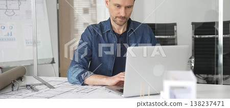 Male architect concentrating on design project using laptop in modern office, surrounded by blueprints, architectural models, and planning tools on the desk Male architect concentrating on design project using laptop in modern office, surrounded by blueprints, architectural models, and planning tools on the desk 138237471