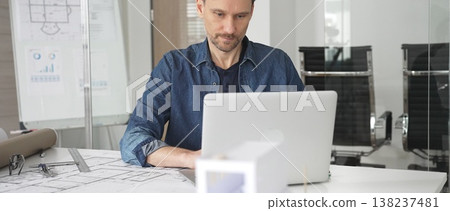 Male architect wearing denim shirt working on laptop in modern office, analyzing blueprints and architectural plans on desk, designing new building construction project 138237481
