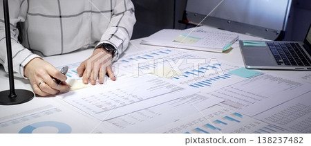Financial advisor examining business reports and charts, analyzing data, planning investments, and preparing forecasts on a cluttered office desk in a late night work environment Financial advisor examining business reports and charts, analyzing data, planning investments, and preparing forecasts on a cluttered office desk in a late night work environment 138237482