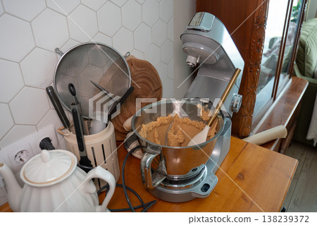 Stand mixer with dough in metal bowl on wooden countertop, kitchen utensils and kettle near tiled wall, home baking preparation scene. Cooking content, recipe illustration, kitchen workflow Stand mixer with dough in metal bowl on wooden countertop, kitchen utensils and kettle near tiled wall, home baking preparation scene. Cooking content, recipe illustration, kitchen workflow 138239372
