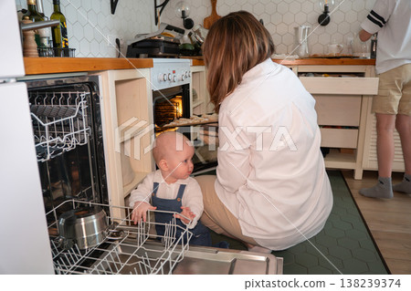 Mother cooks at oven while baby explores open dishwasher in family kitchen, everyday home routine and childcare moment. Parenting, family life, domestic lifestyle, home Mother cooks at oven while baby explores open dishwasher in family kitchen, everyday home routine and childcare moment. Parenting, family life, domestic lifestyle, home 138239374