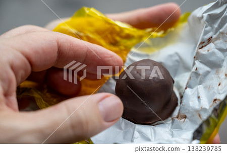 Hand unwrapping cuneo rum chocolate praline, revealing dark, foil-wrapped truffle in yellow wrapper held between fingers, ready to enjoy its bittersweet, boozy flavor 138239785