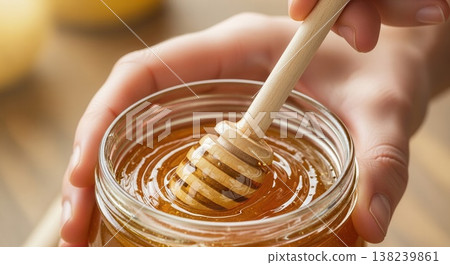 Close-up of a wooden dipper swirling in a jar of golden organic honey, held by hands in warm sunlight. Close-up of a wooden dipper swirling in a jar of golden organic honey, held by hands in warm sunlight. 138239861