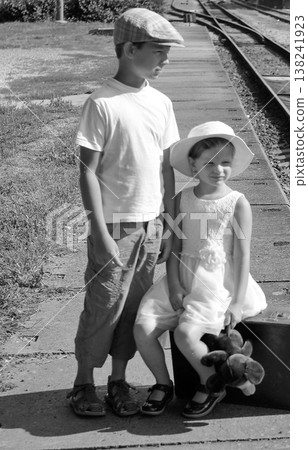 Two children in vintage clothing wait on a train platform with a suitcase and teddy bear 138241923