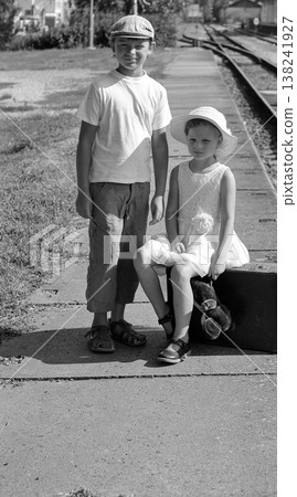 Two children, a boy and a girl, wait on a train platform with a suitcase and teddy bear 138241927