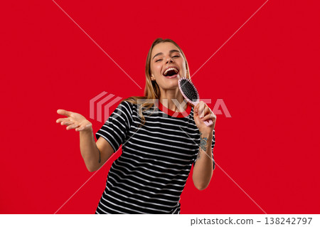 Playful Young Woman Singing and Dancing With Hairbrush On Red Studio Background Playful Young Woman Singing and Dancing With Hairbrush On Red Studio Background 138242797