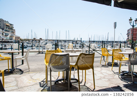 Chairs and tables line a restaurant by the harbor under the bright sunshine on the French island of Corsica in the Mediterranean Sea. 138242856