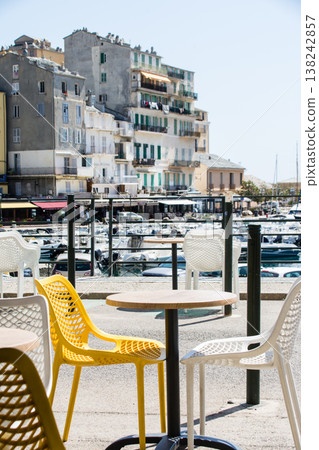 Chairs and tables line a restaurant by the harbor under the bright sunshine on the French island of Corsica in the Mediterranean Sea. 138242857