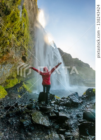 Visitor stands with arms raised in excitement behind Seljalandsfoss Waterfall in Iceland. The sun sets, illuminating the lush landscape and cascading water. Visitor stands with arms raised in excitement behind Seljalandsfoss Waterfall in Iceland. The sun sets, illuminating the lush landscape and cascading water. 138243424