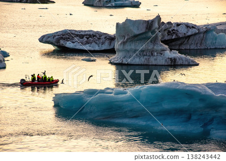 A group of tourists on a boat navigates through a glacier lagoon filled with icebergs. The golden hues of sunset reflect on the water, creating a stunning backdrop A group of tourists on a boat navigates through a glacier lagoon filled with icebergs. The golden hues of sunset reflect on the water, creating a stunning backdrop 138243442