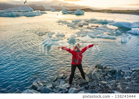 A person stands with arms raised by the ice lagoon in Iceland, celebrating the natural beauty at sunset. Icebergs float in the water, with a colorful sky overhead. 138243444