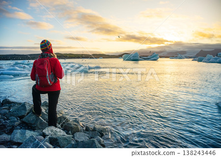 Woman in red jacket joyfully poses on rocky shore of a glacier lake in Iceland. Smiling broadly, she spreads her arms wide, capturing the beauty of the glowing sunset 138243446